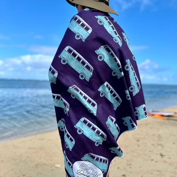 Person wearing a purple towel with teal combie patterns on a beach.